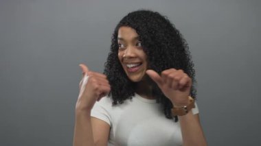 Kıvırcık saçlı kadın gri stüdyoda geniş bir şekilde gülümsüyor ve parmaklarını omzunun üstünden geçiriyor..