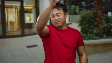 Kırmızı gömlekli adam yeşil ve modern mimari geçmişi olan şehir ortamında güler yüzlü parmak tabancası hareketi yapıyor..