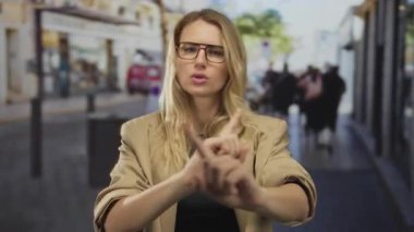 Kadın, şehir merkezinde gözlük ve bej ceket giyerken, açık havada şehir ortamını onaylamadığını belirtirken, hareketli şehir caddesinde parmaklarıyla hareket etmez..