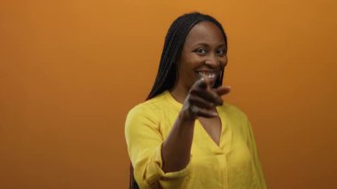 Kadın, turuncu arka plana karşı telefon hareketi yapıyor, neşe ve bağlantı gösteriyor, sarı, kıvırcık saçlar giyiyor, ve izole bir ortamda gülümseyen siyah kadın..