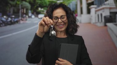 Sokakta elinde tablet ve anahtarla duran kadın, dışarıda emlak ya da iş ortamını temsil ediyor, orta yaşlı, İspanyol kökenli ve profesyonel giyimli..