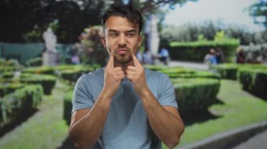 Mavi tişörtlü genç İspanyol adam canlı yeşil bir parkta jestler yapıyor. Etrafı bereketli manzaralarla çevrili, neşeli ve anlamlı bir tavır sergiliyor ve doğal ortamın tadını çıkarıyor..