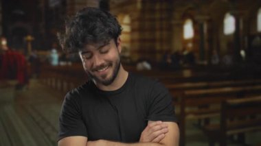 Sıradan giysiler içinde sakallı genç İspanyol bir adam kollarını kavuşturup duruyor loş aydınlatılmış bir kilisede ahşap sıralar ve vitray pencereler var..
