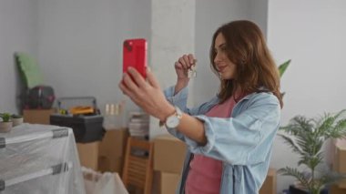 Gülümseyen kadın anahtarlarını tutuyor taşınan kutularla dolu yeni bir dairede selfie çekiyor bitkiler ve akıllı telefonlar ev sahipliğinin mutlu anlarını yakalıyor..