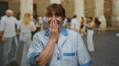 Şehrin kalabalık caddesinde, insanlarla çevrili bir yerde üniforma giymiş bir kadın şaşkınlıkla ağzını kapatıyor..