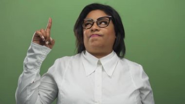 Genç Latin bir kadın yeşil bir duvarın önünde tiksindirici bir ifadeyle yukarıya doğru işaret ediyor..