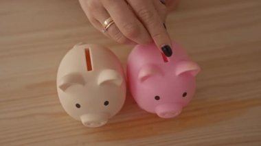 Kadınların elleri, tasarruf, finans, hibe, yatırım, para yönetimi, planlama ve kişisel finans stratejilerini sembolize ederek, tahtadan bir masanın üzerindeki pembe ve bej kumbaralara para koyuyor..