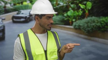 Şehir caddesinde park halindeki arabayı gösteren yüksek görüşlü yelek ve beyaz şapkalı bir adam var..