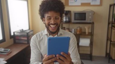 Ofiste gülümseyen Afrikalı Amerikalı bir adam, elinde tablet, hesap makinesi ve doküman gibi ofis esaslarıyla çevrili, çalışma ortamını aktarıyor..