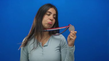 Latin kökenli genç bir kadın bronz madalya tutuyor ve mavi bir stüdyoda baş parmak hareketi yapıyor. Hayal kırıklığı..