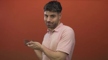 Portakal rengi stüdyo resmiyle ellerini sıvazlayan genç İspanyol adam pembe polo gömlek giyiyor ve nazik bir gülümseme sergiliyor..