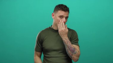 Yüzüne dokunan dövmeleri olan genç bir adam yeşil bir arkaplana dokunuyor. İlgili ifadeler takınıyor. Kişisel bakım ve cilt farkındalığı gösteriyor.