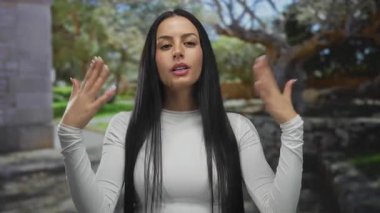 Parktaki genç İspanyol kadın güneşli bir günde kendini yelpazeliyor, yemyeşil ile çevrili ve günlük beyaz bir gömlek giyiyor, açık havanın tadını çıkarıyor.