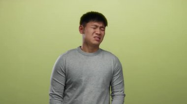 Gri kazaklı genç Çinli adam, izole edilmiş sarı bir arka plana karşı rahatsızlıkla tepki veriyor ve etkileyici vücut dilini vurguluyor..