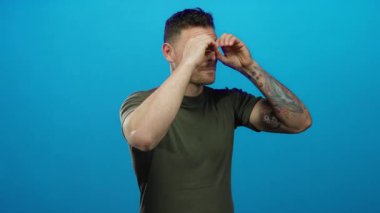 Dövmeli genç adam, neşeli mavi arka plana karşı dürbünlü el hareketi yapıyor. Oyunculuğu ve merakı etkileyici bir şekilde gösteriyor..
