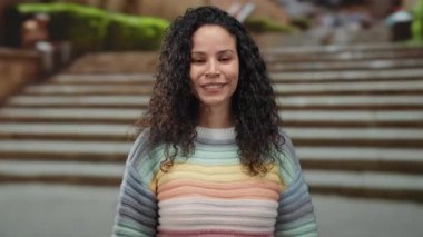 Yüzlerini dışarıda yapan, renkli kazak giyen, basamaklarda duran, kıvırcık saçlı, yetişkin İspanyol kadın, sokak manzaralı, neşeli şehir ortamı.