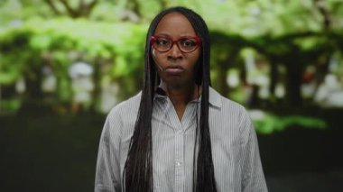 Parkta kıvırcık saçlı bir kadın jestler yoluyla ifade ediyor, kırmızı gözlük takıyor, duygularını açık havada iletiyor, canlı yeşil bir ortamda doğal güzelliğini sergiliyor..