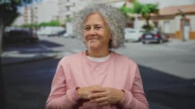 Güneşli bir günde, şehir caddesinde gri kıvırcık saçlı, pembe kazaklı, sıcacık gülümseyen bir kadın..