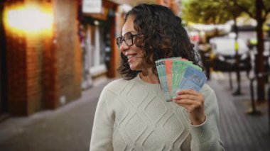 Orta yaşlı bir kadın elinde Malezya bankası, canlı bir şehir caddesinde duruyor, neşeli bir ifade sergiliyor..