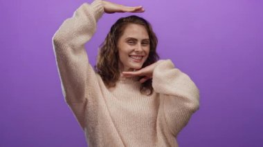 Rahat süveter içinde gülümseyen kadın, canlı mor arka plana karşı çerçeveleme hareketi yapıyor, yaratıcılık ve neşe ifade ediyor..