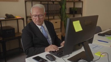 Modern ofis içinde akıllı telefon ve bilgisayar kullanan kıdemli bir adam. İşyerinde raflar ve temiz bir masa ile odaklanmış ve meşgul görünüyor..