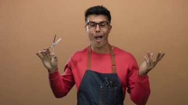 Gözlüklü ve önlüklü genç bir adam kahverengi bir arka plana karşı makas tutuyor ve berberlik mesleğini gösteriyor..