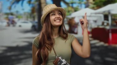Piyasada gülümseyen kadın, başparmağıyla yukarı doğru bakıyor. Canlı bir sokak ortamında kamera ve güneş şapkasıyla neşeli turizm sergiliyor..