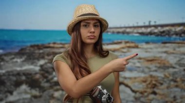 Deniz kenarında ciddi bir ifadeye sahip bir kadın, bir şapka takıyor ve bir kamera tutuyor, manzaralı bir kayalık plaj ve mavi okyanus arka planına karşı kurulmuş, seyahat ve keşifleri vurguluyor.