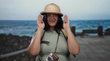 Sahilde gezen, şapkalı, sırt çantalı ve kameralı gülümseyen bir kadın deniz manzaralı arka planda seyahat macerasının tadını çıkarıyor..