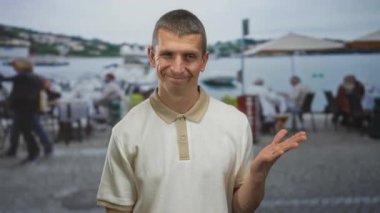 Açık hava restoranının terasında masalarla gülümseyen bir adam ve arka planda rıhtım manzarası yakınında insanlar bulanıklaştı..