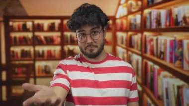 Genç, sakallı, gözlüklü ve çizgili tişörtlü İspanyol adam, kapalı alanda entellektüel bir atmosfer yaratan renkli kitaplarla çevrili bir kütüphanede güvenle duruyor..