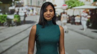 Güneşli caddede gülümseyen bir kadın yeşil elbiseli, açık hava kafeleri ve bulanık yürüyen insanlarla dolu şehir manzaralı bir yerde güven içinde yol gösteriyor..