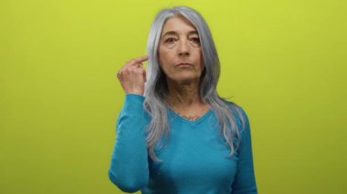 Yaşlı gri saçlı kadın müstehcen bir jest yapıyor. İki orta parmağı da havada, parlak sarı arka plan duvarına karşı duruyor. Sert bir ifadeyle..