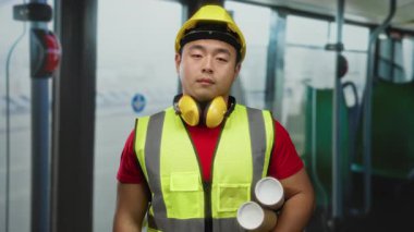 Güvenlik yeleği ve kasketi takmış, şehir gelişimini ve toplu taşıma planlarını sembolize eden Asyalı genç bir adam otobüsün içinde duruyor..
