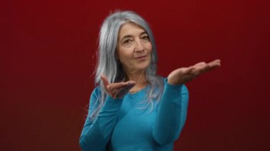 Gri saçlı, mavi kazaklı yaşlı kadın elini kırmızı duvara uzatıyor ve izole bir stüdyo çekiminde dostane bir gülümseme sergiliyor..