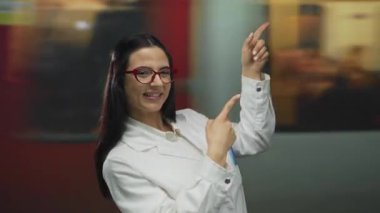 Kırmızı gözlüklü kadın kapalı mekanda durup bulanık tren istasyonunu işaret ediyor..