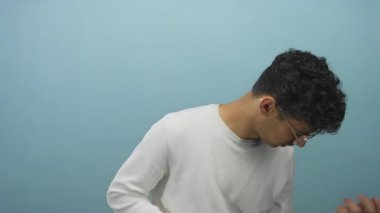 Beyaz gömlekli ve gözlüklü İspanyol adam başını öne eğiyor. Stüdyoda mavi duvara karşı sağ elini inceliyor..