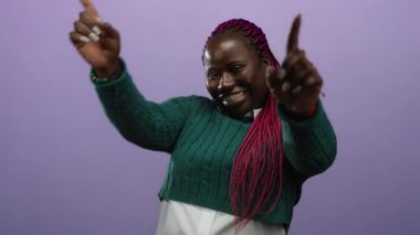 Canlı kırmızı örgüleri ve yeşil süveteri ile mor izole edilmiş arka planda güvenle gülümseyen kadın kameraya neşeli bir şekilde bakıyor..