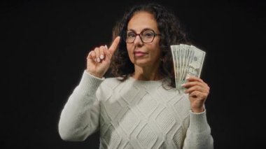 Amerikan dolarlarını elinde tutan kadın şaşırmış bir ifadeyle siyah bir arkaplanı işaret ediyor, merak ve finansal planlama sergiliyor..