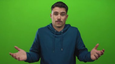 Gündelik kıyafetli genç adam yeşil bir arka planın önünde duruyor. Kaldırılmış elleri ve belirsiz bir ifadesi var. Bir anlık şaşkınlığı yakalıyor..