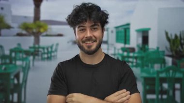 Sakallı genç adam neşeli bir şekilde açık havada canlı bir restoran terasında güneşli bir gökyüzünün altında canlı yeşil sandalyelerle geziyor..