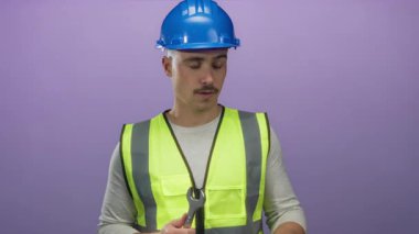 Yansıtıcı yelek ve şapka giyen genç İspanyol bir adam mor arka plan duvarına İngiliz anahtarı tutarken endişeli görünüyor..