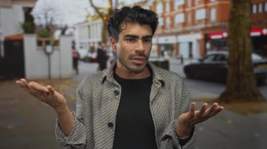 Erkek, çatık kaşlarıyla omuz silkerken bir sokak ortamında göğsünün önünde ellerini açık tutar; hayal kırıklığı.