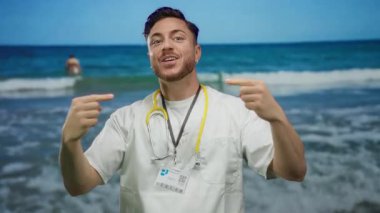 Sakallı ve steteskoplu genç doktor iki eliyle kendini gösteriyor, deniz kenarında duruyor ve arka planda dalgalar var..