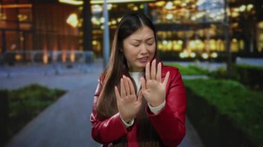 Şehirde açık havada kırmızı ceket giyen kadın, akşam ışıklarını ve modern mimariyi tasvir eden bulanık şehir arka planına karşı el hareketleriyle reddediyor..