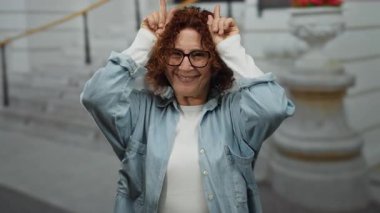 Esmer, orta yaşlı, gülümseyen, parmaklarıyla boynuz hareketi yapan şehir sokaklarında gözlük takan, arka planda bulanık şehir yaşam tarzını gösteren bir kadın.