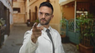 İspanyol gözlüklü ve beyaz önlüklü bir adam eski bir şehir caddesinde ciddi bir ifadeyi işaret ederek şehir ortamında profesyonel ya da otoriter bir kişiliği işaret ediyor..