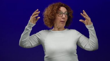 Mavi arka plan karşısında esprili bir şekilde el kol hareketi yapan gözlüklü ve beyaz gömlekli bir kadın izole ortamda eğlenceli duygular taşıyor.