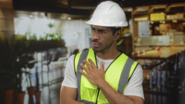 Kask takan ve yelek giyen adam ellerini göğsüne koyuyor ve inşaatta endişeli görünüyor; sorumluluk kaygısı.