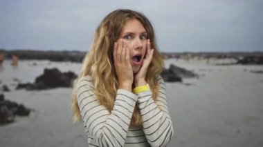 Kadın kumsalda ellerini kaldırır genç sarı saçlarını sergilerken ağzını geniş gözlerle çizgili gömlekle kapatır..